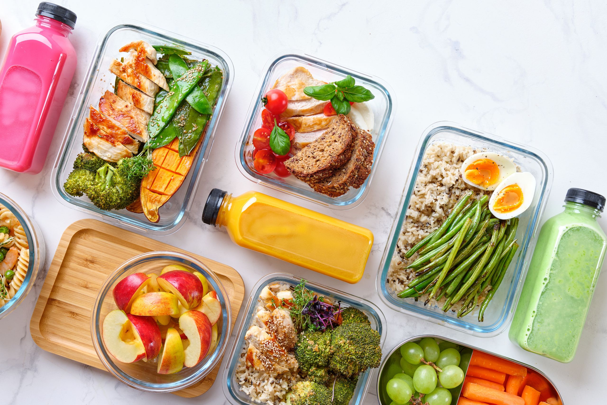 Colorful meal prep containers with healthy food including grilled chicken, vegetables, rice, eggs, fruit, and smoothies on a white kitchen counter.