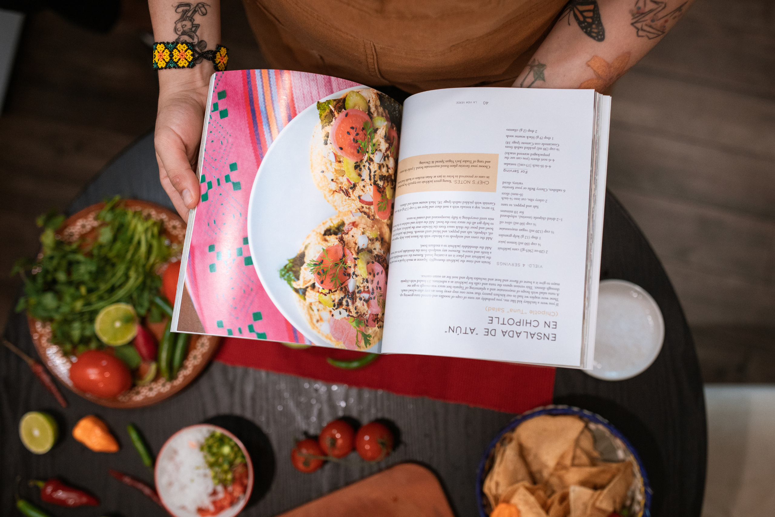 Person holding an open spice cookbook above a kitchen table with fresh vegetables, spices, and ingredients.