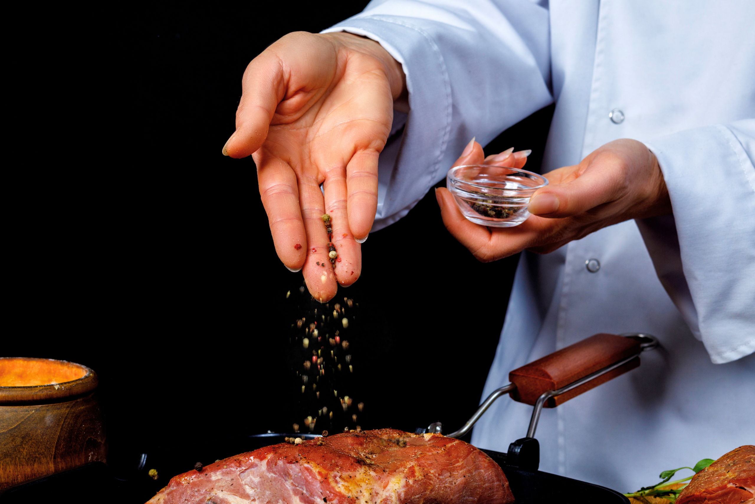 A close-up shot of a chef's hands, one hand sprinkling a mixture of colorful peppercorns over a large cut of raw red meat, and the other holding a small glass bowl containing more of the spice blend. The chef is wearing a white coat.