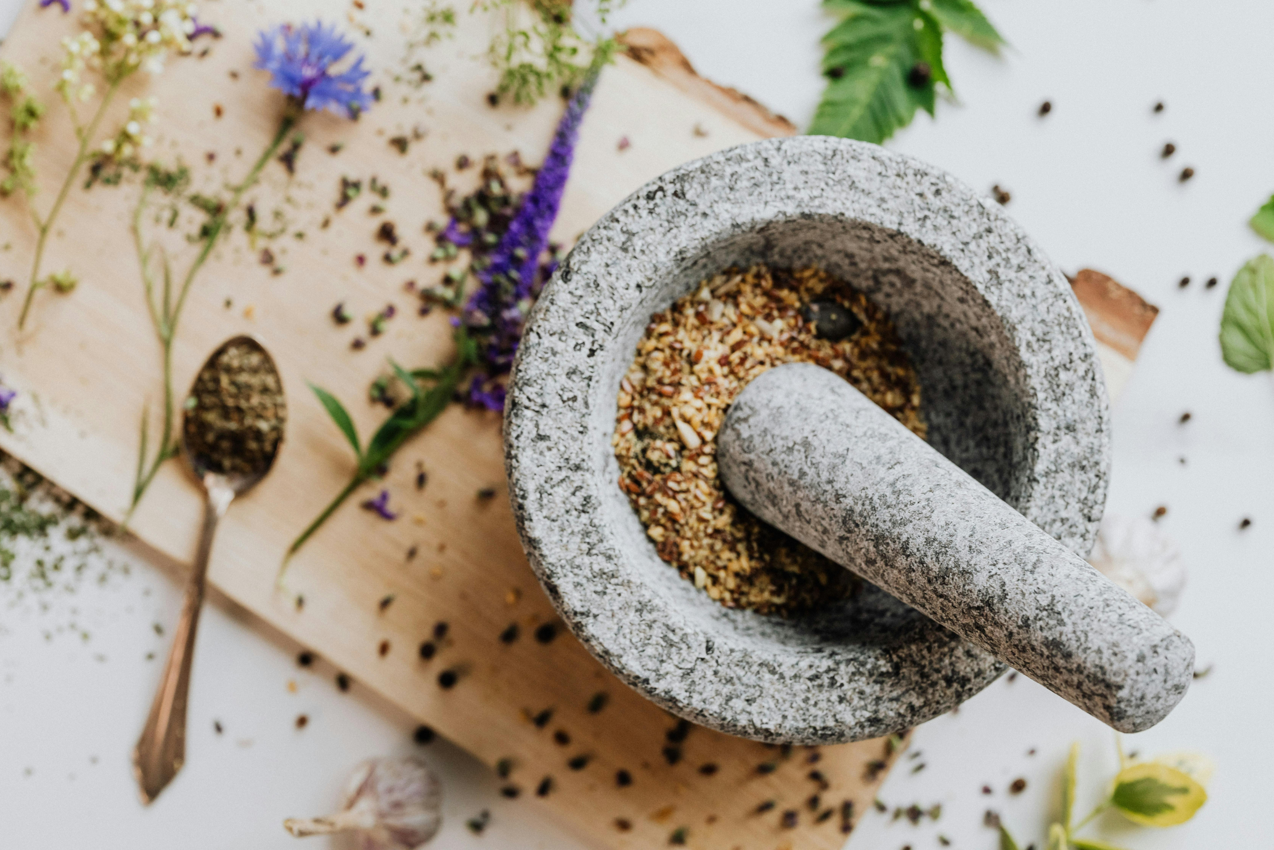 Stone mortar and pestle filled with ground spices on a wooden board surrounded by scattered herbs and dried flowers.