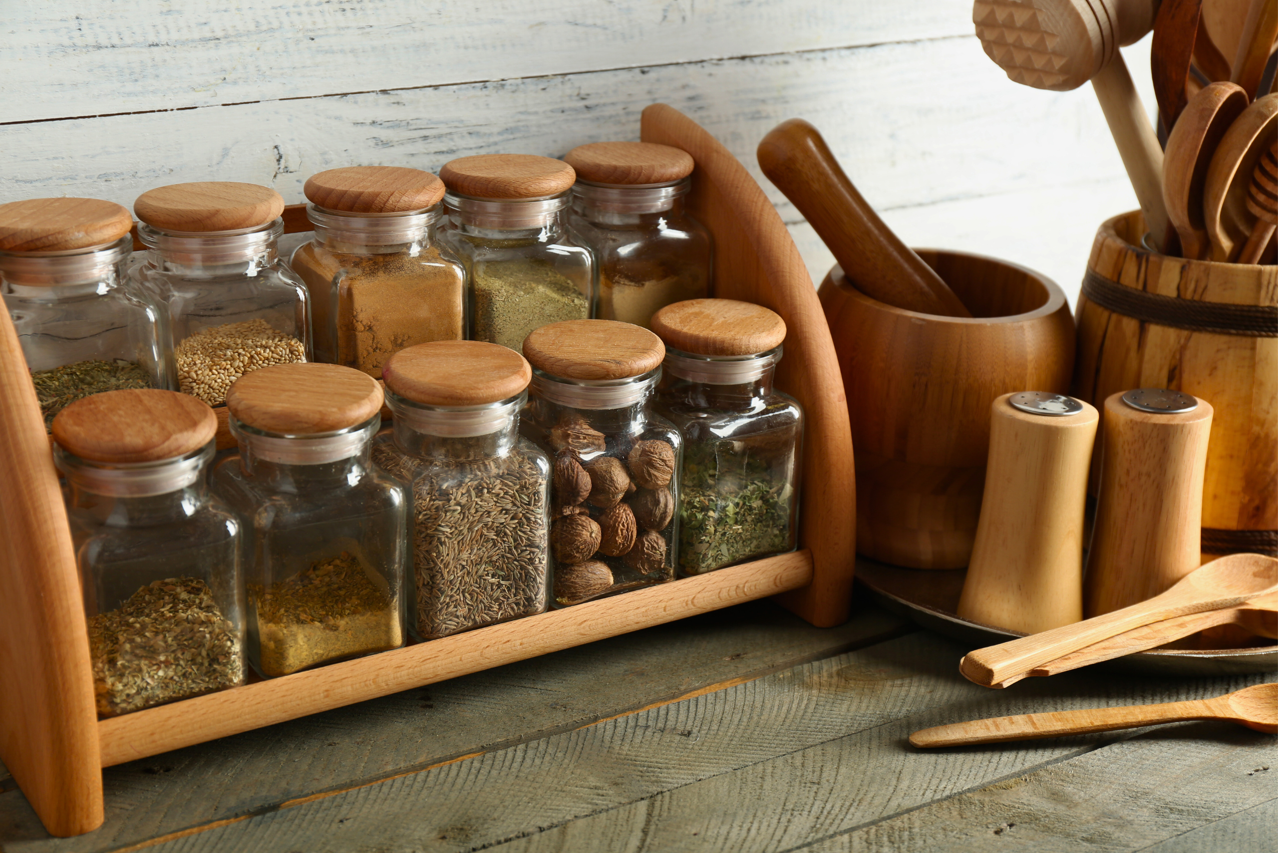 Wooden spice rack holding glass jars filled with herbs and spices, placed on a rustic kitchen countertop beside wooden cooking utensils.