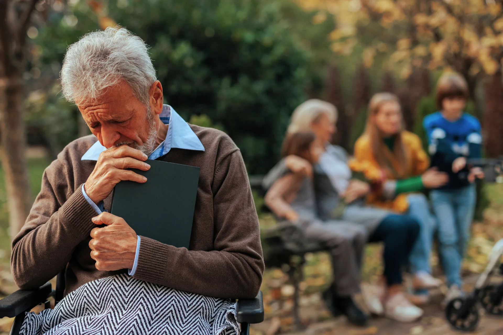 An elderly man sits alone on a bench holding a closed book, appearing emotional, while blurred family members talk in the background — symbolizing generational distance and emotional abandonment.