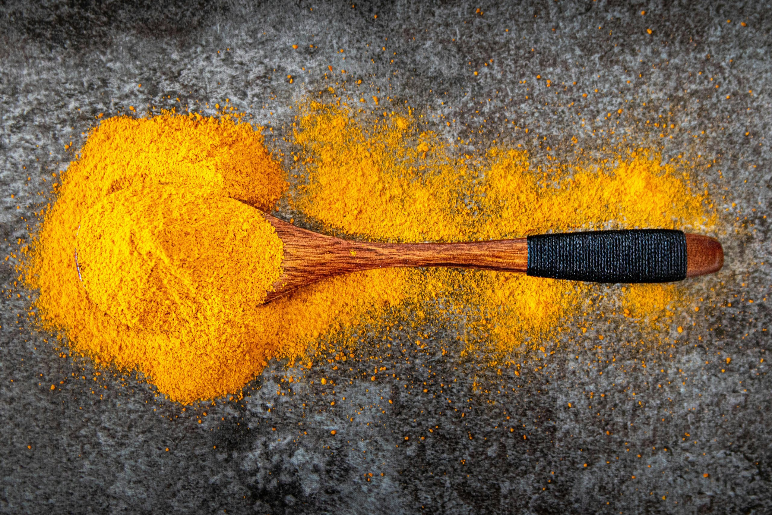 Wooden spoon filled with vibrant yellow homemade curry powder spilled on a dark stone surface.