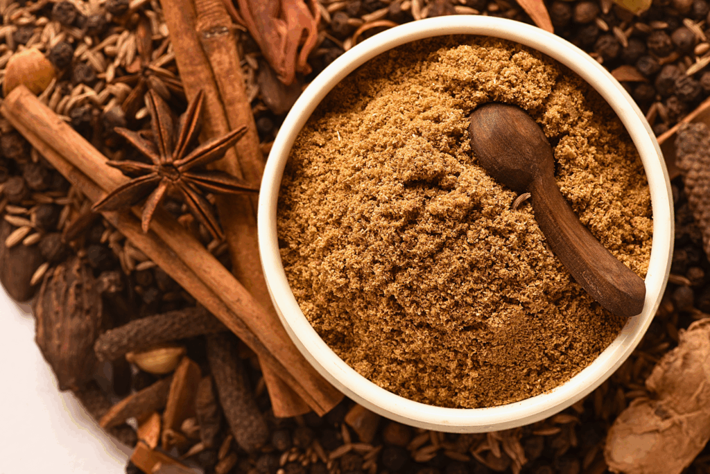 “Bowl of freshly ground garam masala spice blend with a wooden spoon, surrounded by whole spices like cinnamon, star anise, cumin seeds, and cloves