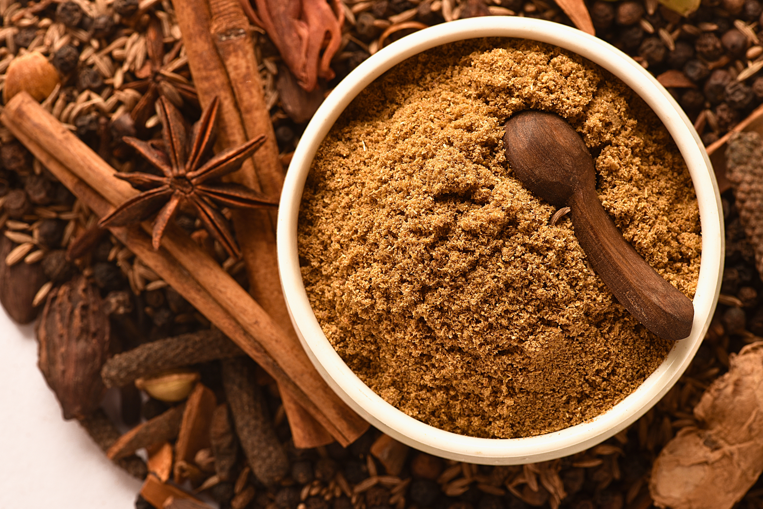 “Bowl of freshly ground garam masala spice blend with a wooden spoon, surrounded by whole spices like cinnamon, star anise, cumin seeds, and cloves